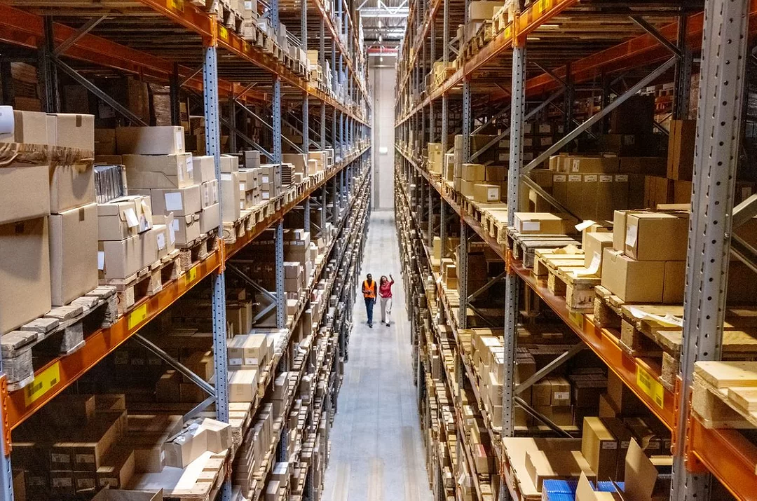 A MAN AND A WOMAN IN HARD HATS WALK DOWN A VAST WAREHOUSE AISLE, SEEN FROM ABOVE