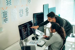 TWO PEOPLE AT A DESK CONSULT OVER A COMPUTER SCREEN