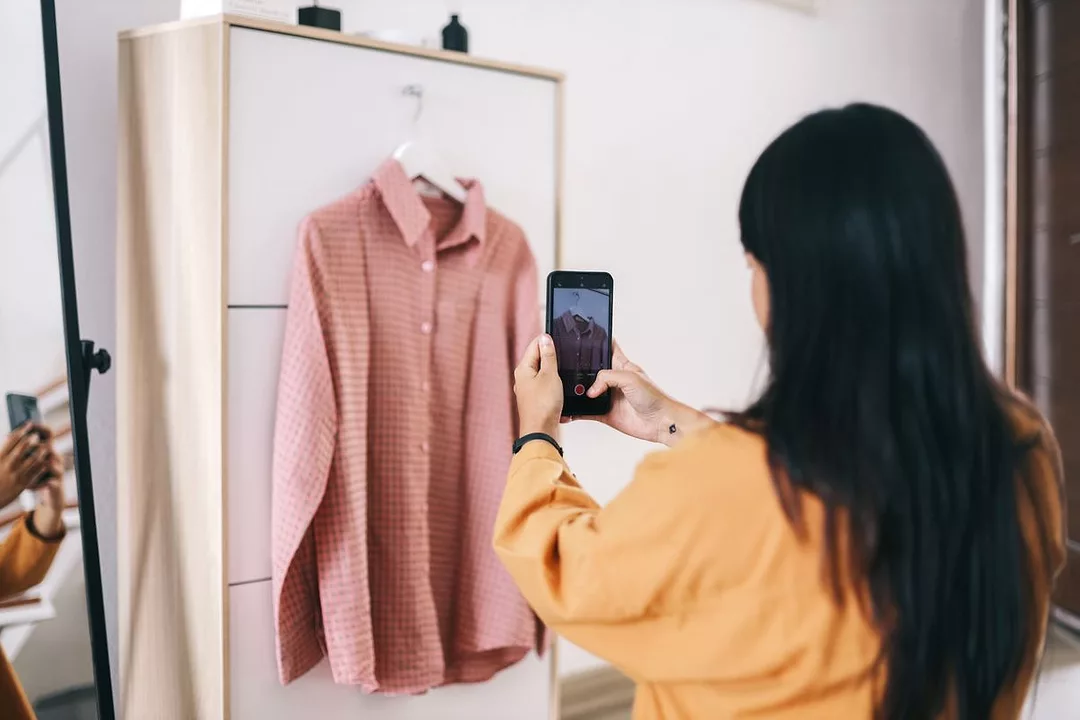 A WOMAN PHOTOGRAPHS A SHIRT ON A HANGER WITH HER PHONE