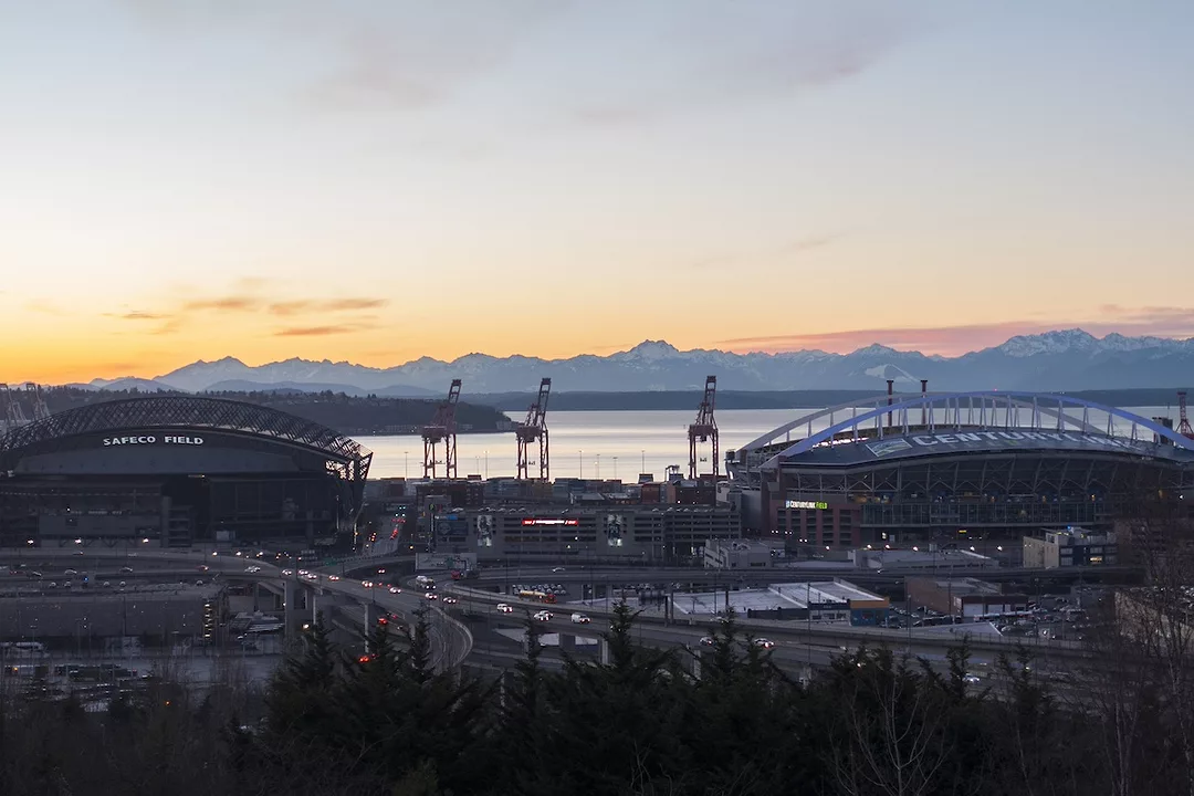Sunset of a cityscape along a large body of water, with two sports stadiums positioned on the left and right, and a pair of orange shipping cranes along the shore in the background
