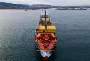 A red container ship stacked with yellow shipping containers, moving through a large expanse of sea with a distant shoreline in the background