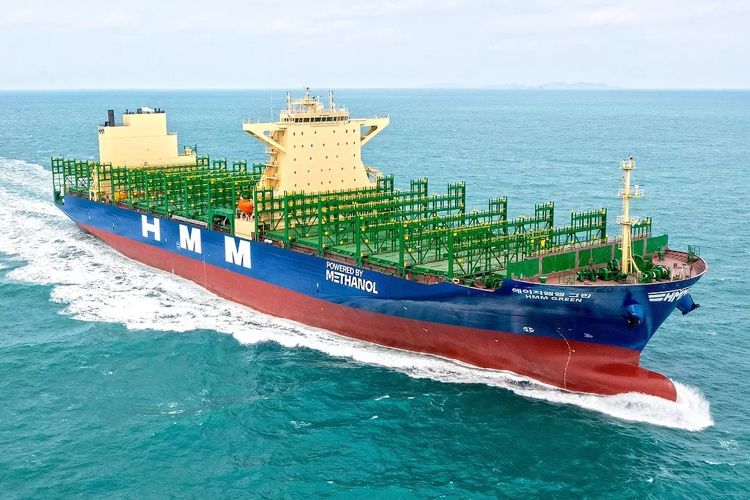 A blue and red striped container ship with "HMM" written along the side in large white lettering, with a green deck and two yellow towers