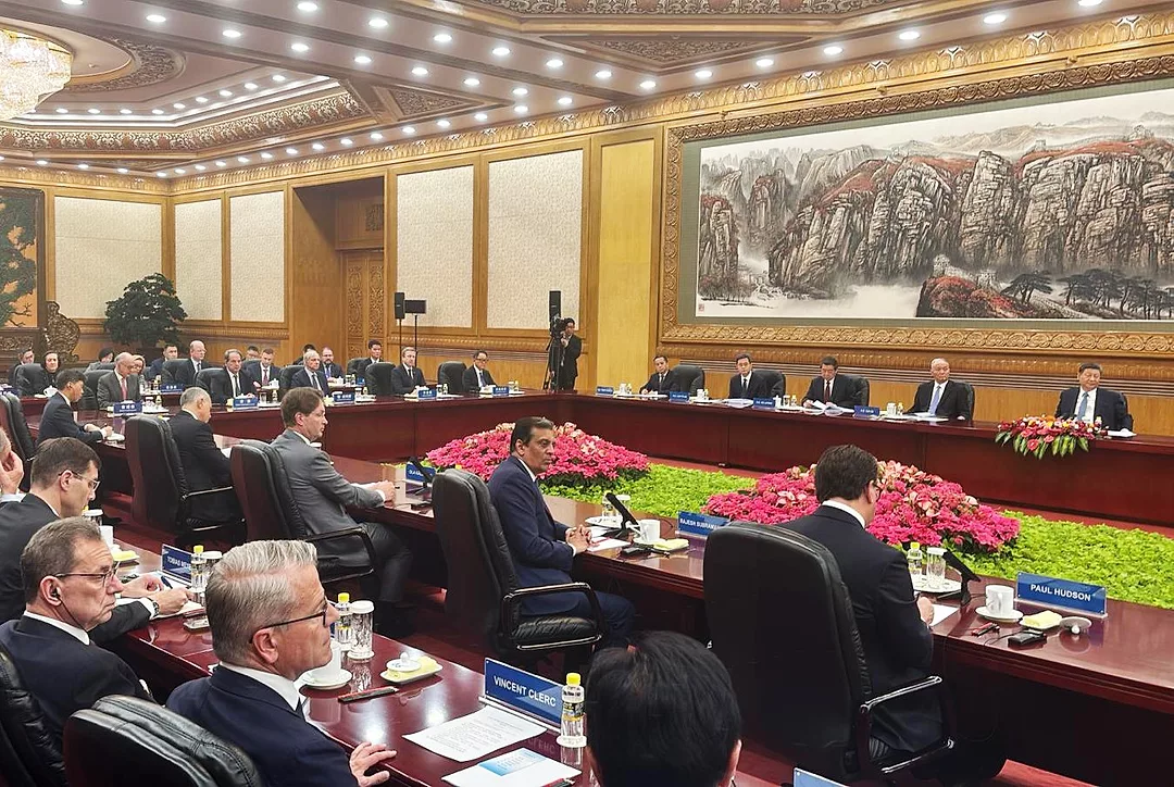 IN A LARGE GILDED CONFERENCE ROOM, DOZEN OF MEN IN SUITS LISTEN TO A BANK OF MEN SITTING ON A DAIS