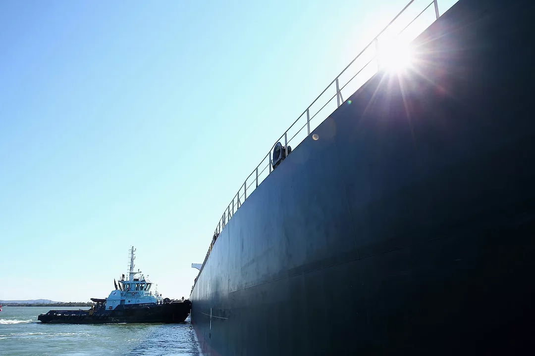 A close-up of the hull of a large black container ship at sea, with another black ship in the background