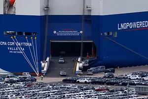 DOZENS OF CARS SIT ON A DOCK, WITH SOME DRIVING INTO THE MOUTH OF A COMMERCIAL RORO VESSEL