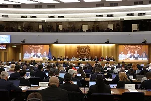 MANY MEN AND A FEW WOMEN SIT IN A GIANT MEETING HALL FACING A FLAT-SCREEN TV WITH MORE PEOPLE ON IT