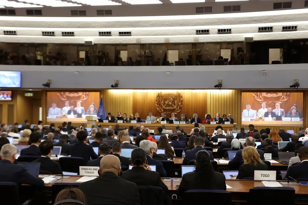 MANY MEN AND A FEW WOMEN SIT IN A GIANT MEETING HALL FACING A FLAT-SCREEN TV WITH MORE PEOPLE ON IT