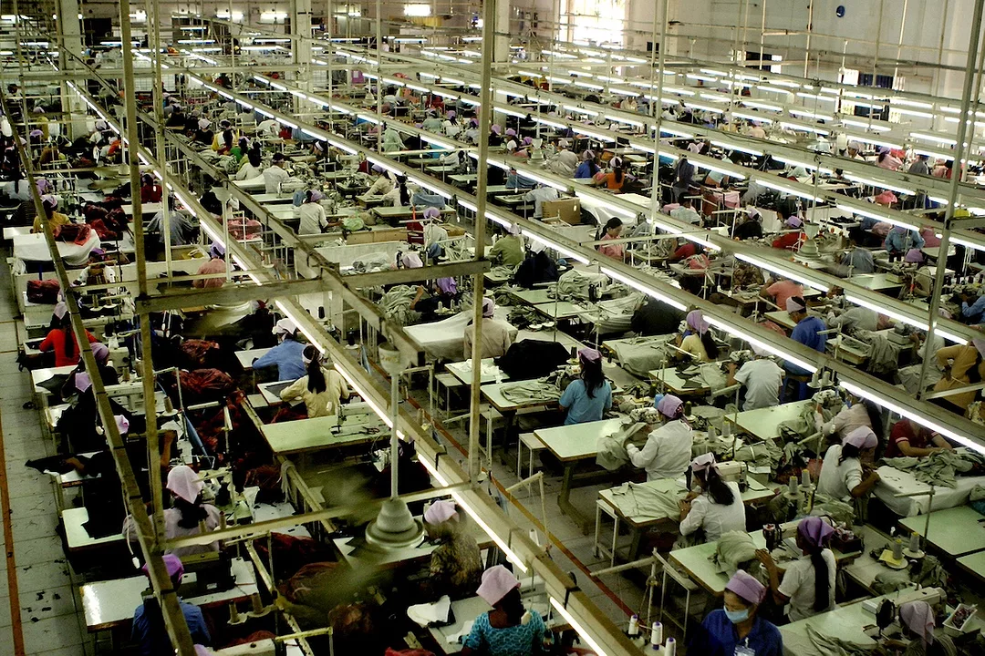 A dimly-lit garment factory floor, filled with rows of workers in blue shirts sewing clothing at white tables