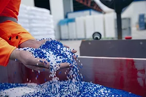 A person wearing an orange jacket scooping a handful of blue and white plastic pellets out of a large red and grey metal box