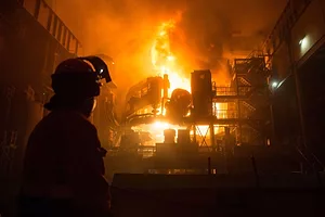 A WORKER IN SILHOUETTE LOOKS ON AT A SCENE OF ENORMOUS FIRES ENGULFING LARGE PIECES OF EQUIPMENT