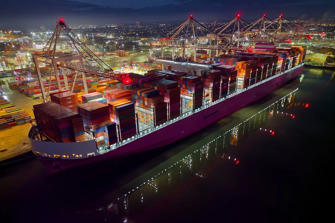 A large purple container ship docked beneath a row of cranes a night, and stacked with multi-colored shipping containers