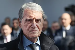 An elderly man with greying hair, wearing a blue tie and black suit jacket