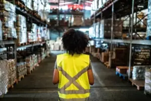 A dark-skinned woman wearing a neon yellow vest with her back to the camera, standing in an aisle flanked by warehouse shelves on either side