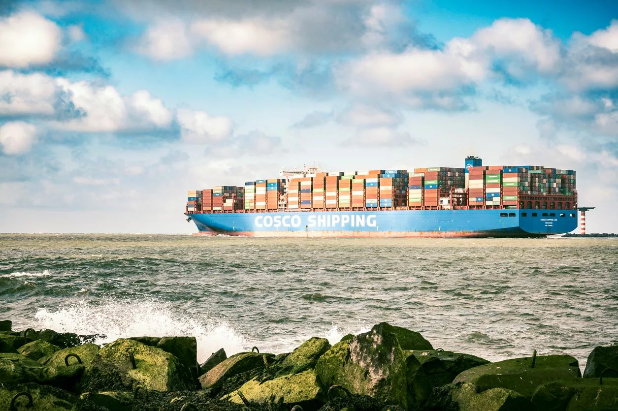 Üzerinde dev beyaz harflerle COSCO yazıyor bir konteyner gemisi okyanusu yarmakta; ön planda kayalar olunuyor