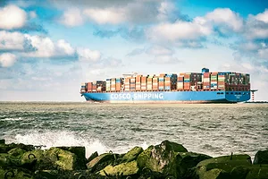 A CONTAINER SHIP BEARING THE WORD COSCO IN GIANT WHITE LETTERS PLIES THE OCEAN, WITH ROCKS IN THE FOREGROUND