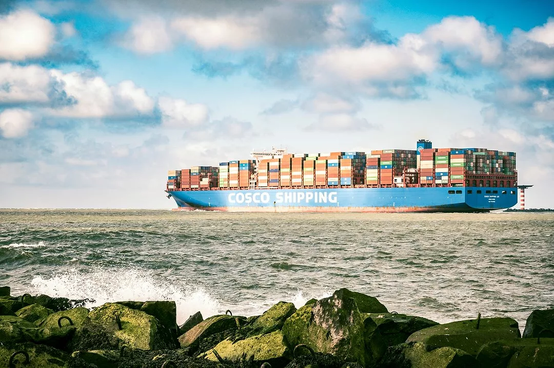 A CONTAINER SHIP BEARING THE WORD COSCO IN GIANT WHITE LETTERS PLIES THE OCEAN, WITH ROCKS IN THE FOREGROUND