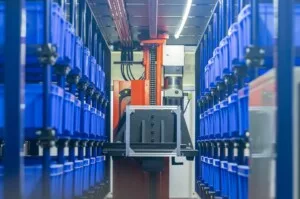 BLUE PLASTIC CRATES SIT IN MULTI-ROW SHELVING IN A WAREHOUSE, A YELLOW ROBOTIC MACHINE VISIBLE AT THE END OF THE AISLE