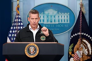 A man in a black windbreaker and white collared shirt standing at a black podium bearing the U.S. presidential seal, with a blue logo of the White House directly behind him