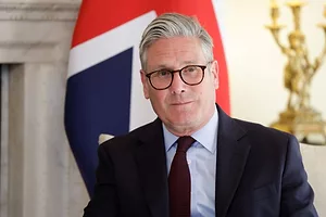 A MAN IN A SUIT WEARING SPECTACLES, WITH GREYING HAIR, SITS IN FRONT OF A UNION JACK FLAG