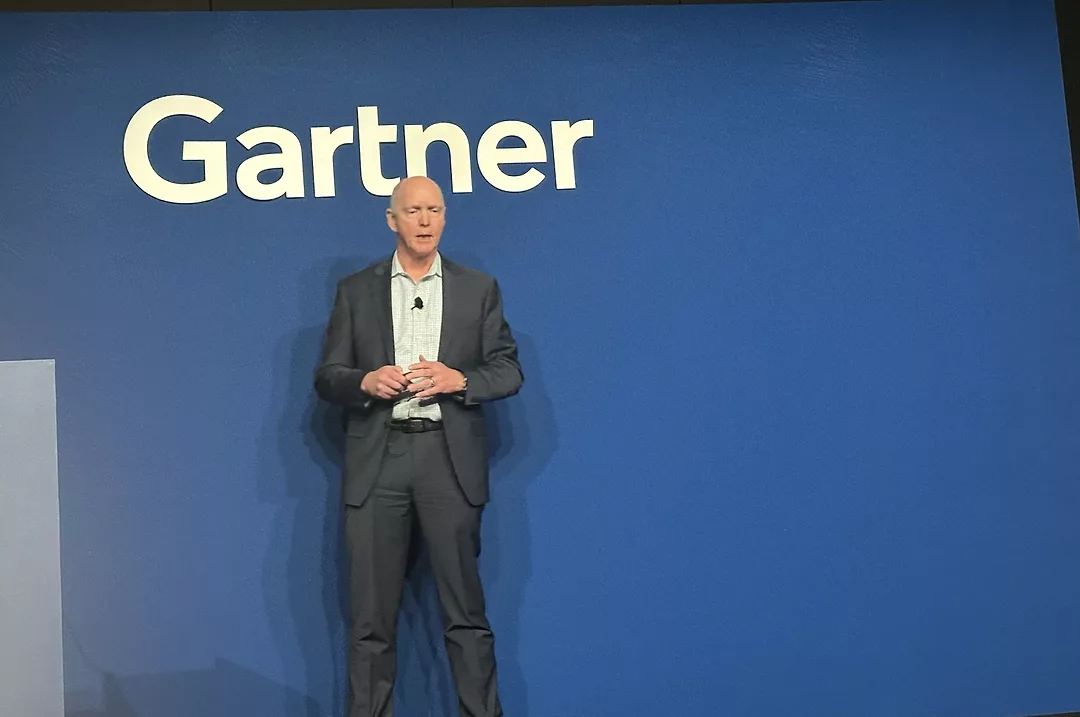 A TALL MAN IN A BLACK SUIT ADDRESSES AN AUDIENCE FROM A STAGE WITH A SIGN THAT SAYS GARTNER