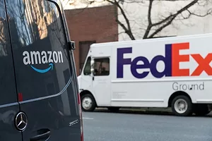 AN AMAZON VAN STANDS PARKED IN A STREET, WITH A FEDEX VAN VISIBLE IN THE DISTANCE