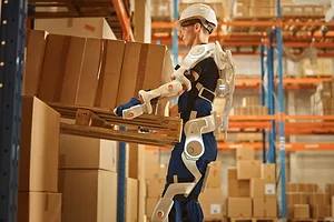 A WORKER WEARING A WHITE ROBOTIC EXOSUIT LIFTS A BOX IN A WAREHOUSE