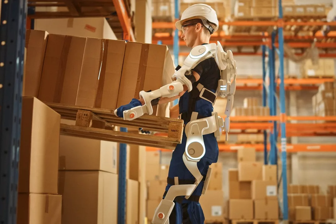 A WORKER WEARING A WHITE ROBOTIC EXOSUIT LIFTS A BOX IN A WAREHOUSE