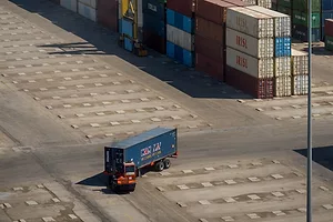 A blue-semi-truck driving out of rows of multi-colored shipping containers in a concrete lot