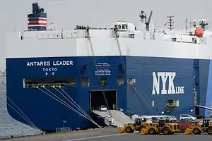 A navy blue container ship with a white deck, docked along a concrete shoreline