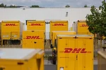 Three rows of yellow box trucks parked in front a white sheet metal building, with "DHL" written in red letter across the back