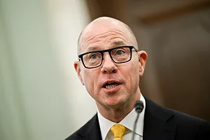 A bald man with black-rimmed glasses, wearing a suit with a yellow tie