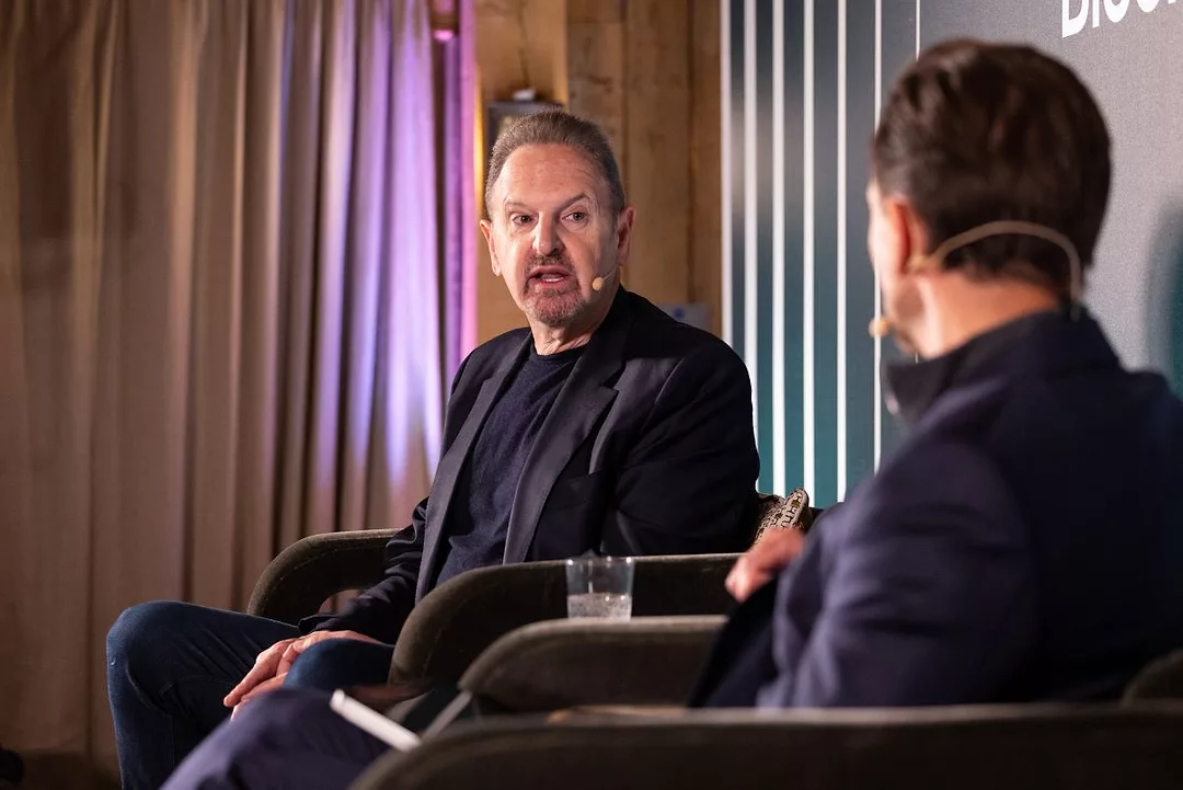 A MAN IN A BLACK SUIT AND TEESHIRT WEARING A HEADSET SITS TALKING AT A CONFERENCE