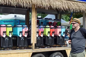 A MAN IN SUNGLASSES WALKS PAST A LONG ROW OF CANDY DISPENSERS IN MULTIPLE FLAVORS AND COLORS, ALL UNDER A GRASS ROOF