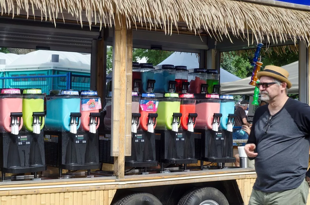 A MAN IN SUNGLASSES WALKS PAST A LONG ROW OF CANDY DISPENSERS IN MULTIPLE FLAVORS AND COLORS, ALL UNDER A GRASS ROOF