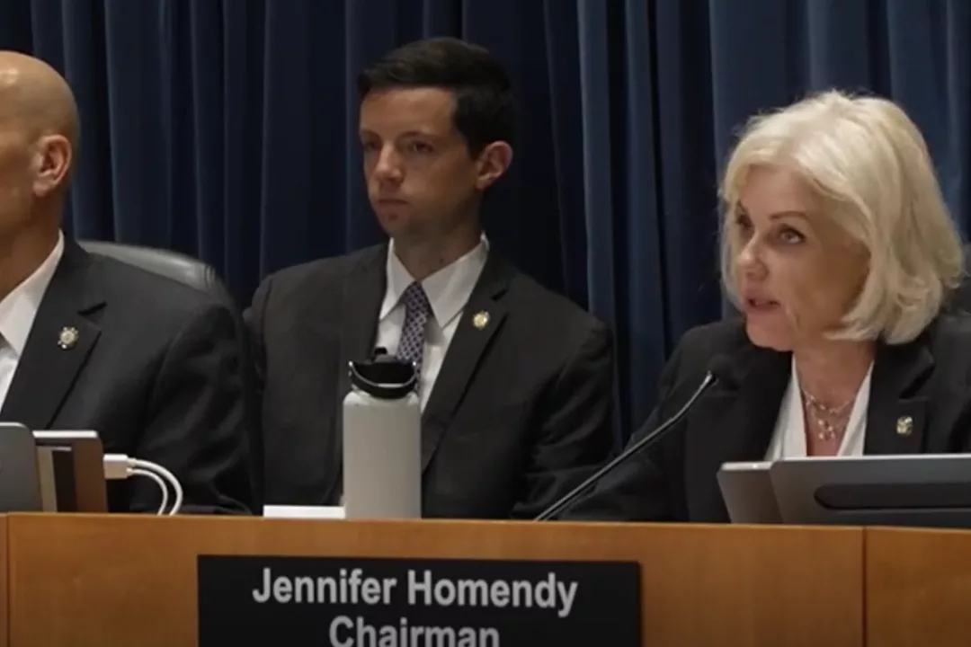 A white-haired woman in a black sport coat and a white dress shirt sitting at a dais in front of a microphone, with two other men in suits sitting to her right