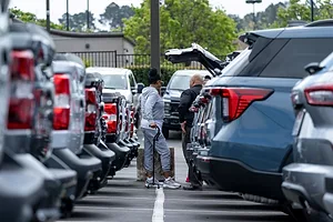 TWO PEOPLE EXAMINE A CAR WITH AN OPEN TRUNK, IN A LONG LINE OF CARS