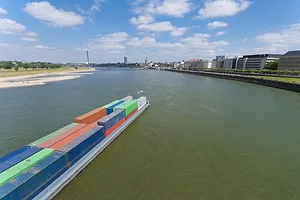 A barge moving through a narrow river flanked by buildings on either side, and carrying rows of multi-colored shipping containers