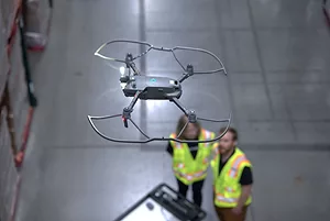 SEEN FROM ABOVE, A DRONE HOVERS IN A WAREHOUSE AISLE, OUT OF FOCUS PEOPLE IN HI VIS VESTS BELOW