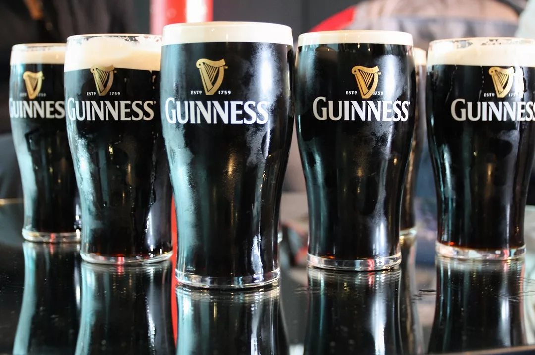 FIVE FULL GLASSES OF DARK BEER WITH IVORY HEADS BEAR THE GUINNESS LOGO WITH A HARP