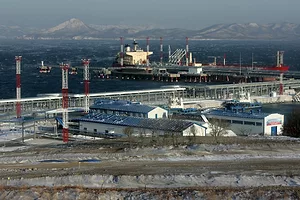 Blue-roofed warehouse buildings in a row along a coastline, with grey shipping docks dotted with red and white cranes in the background, and a mountain range towering over the horizon beyond the waterline