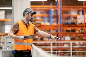 A WORKER IN A HI-VIS VEST WEARING A HEADSET AND HOLDING A TABLET COMPUTER GIVES DIRECTIONS TO WORKERS BELOW