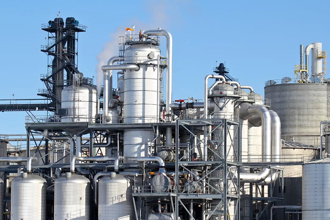 A white factory with rows of silos and pipes and white smoke coming from smokestacks
