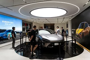 PEOPLE CLUSTER AROUND A SHINY SILVER CAR SITTING ON A ROTATING DISPLAY