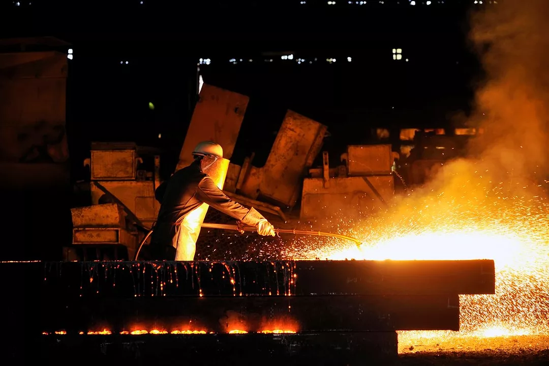 A WORKER IN PROTECTIVE GEAR AND A HEAVY VISOR WIELDS A ROD AMONG DRAMATIC CLOUDS OF SPARKS IN A DARK FACTORY