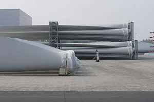 GIANT METAL PARTS SIT ON TARMAC OUTSIDE, AS A WORKER SWEEPS THE GROUND, A LARGE FACTORY BUILDING IN THE BACKGROUND