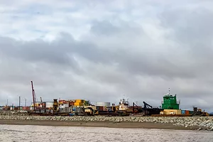 Stacks of multi-colored shipping containers and a tall red crane along a rocky coastline on an overcast day