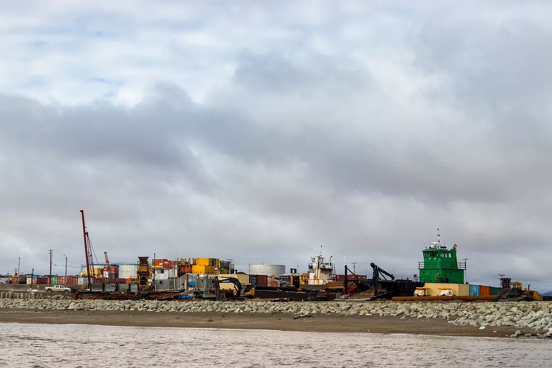 Stacks of multi-colored shipping containers and a tall red crane along a rocky coastline on an overcast day