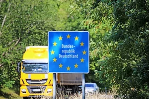A BRIGHT YELLOW FREIGHT TRUCK APPROACHES A BLUE AND WHITE EU BORDER SIGN