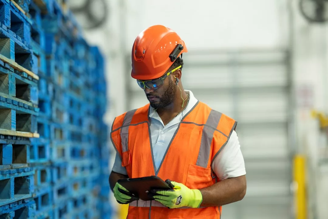 A MAN IN HI-VIS VEST AND BRIGHT ORANGE HELMET LOOKS AT A TABLET COMPUTER AMONG STACKED PALLETS IN A WAREHOUSE