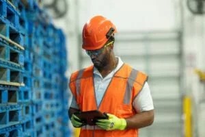 A MAN IN HI-VIS VEST AND BRIGHT ORANGE HELMET LOOKS AT A TABLET COMPUTER AMONG STACKED PALLETS IN A WAREHOUSE